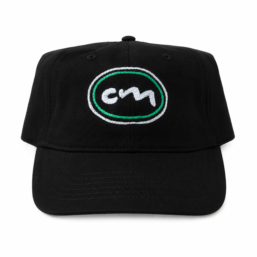 A black baseball cap featuring an embroidered oval logo on the front with a white stylized 'cm' inside a green and white border.