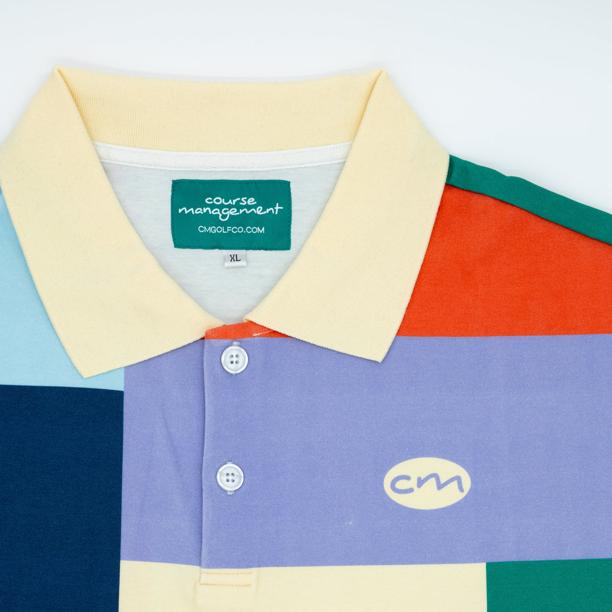 A close-up of a multicolor block-patterned polo shirt by Course Management, featuring a light yellow collar and a color-blocked design of purple, navy, green, orange, and pale yellow.