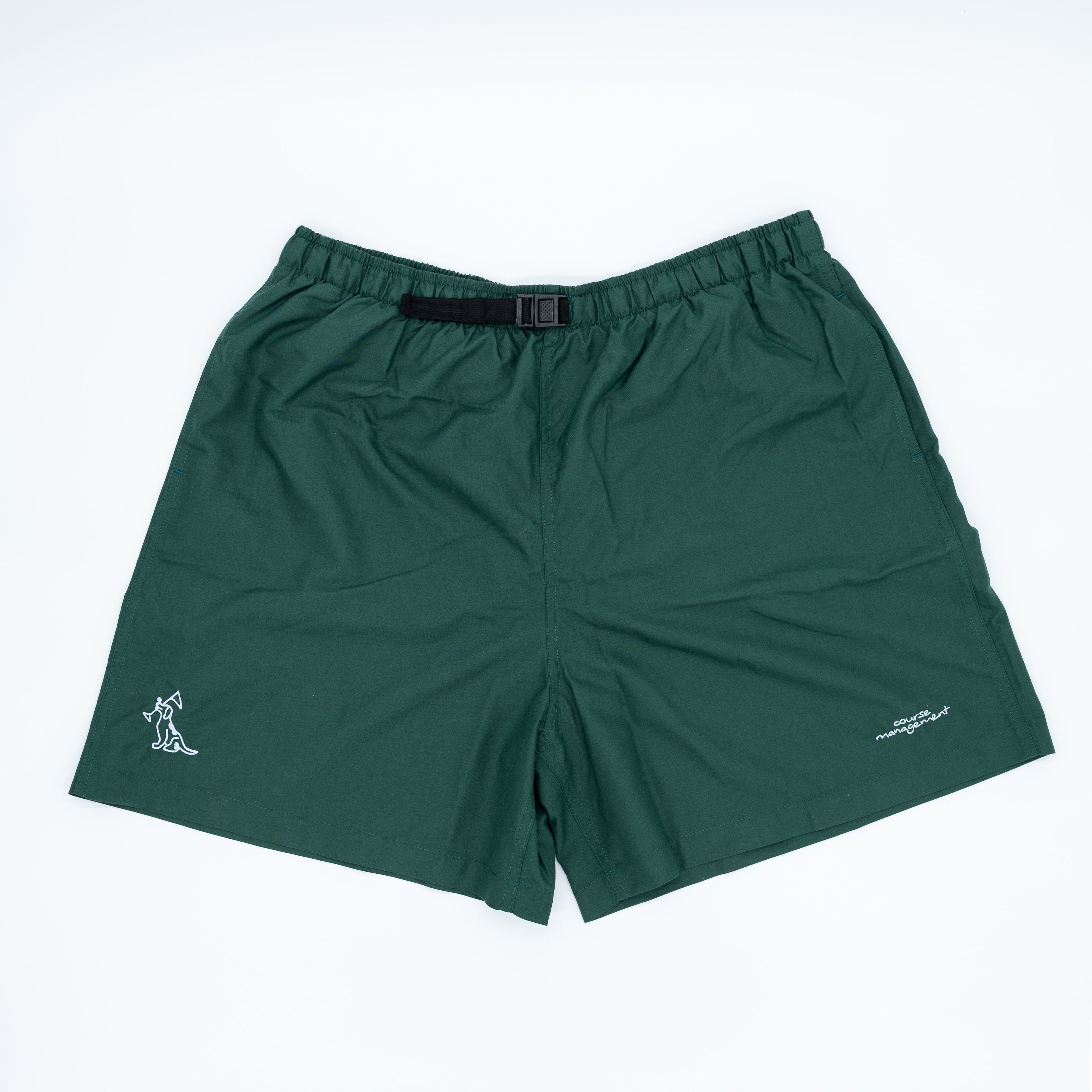 Dark green athletic shorts with an elastic waistband and a black belt, featuring a small white embroidered dog logo on the left leg and a small white script logo on the right leg, displayed flat on a white background.