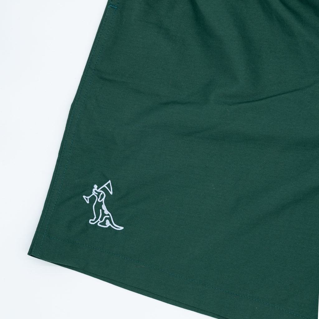 A close-up of a dark green shorts, featuring a small white embroidered dog logo on the lower left hem, shot against a plain white background.