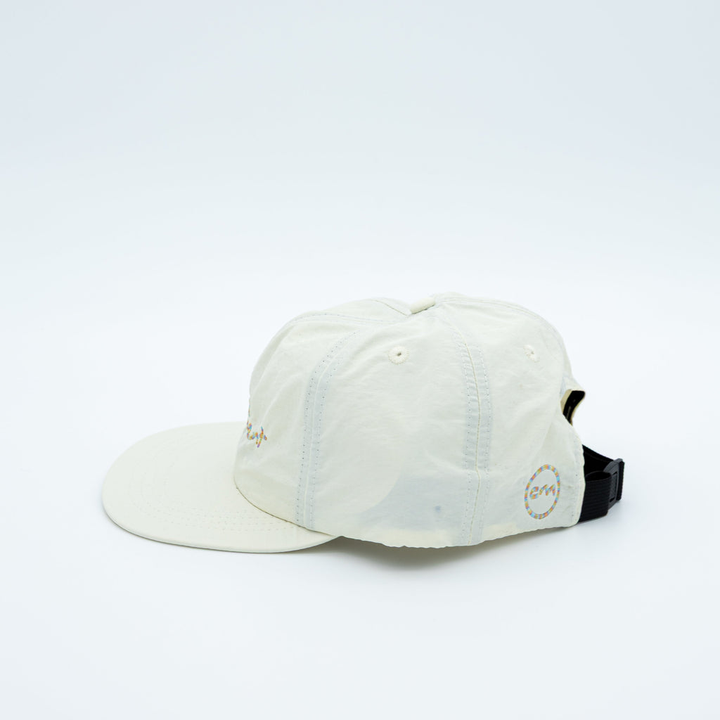 An off-white cotton 5-panel baseball cap with a curved brim, featuring a circular logo patch on the back, shown against a plain white background.