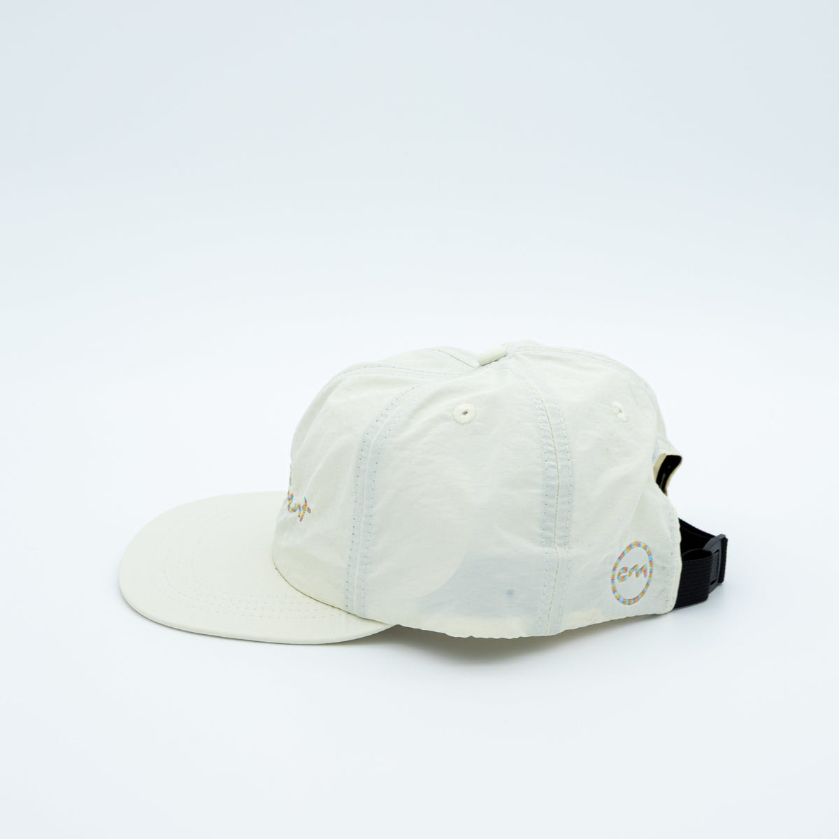 An off-white cotton 5-panel baseball cap with a curved brim, featuring a circular logo patch on the back, shown against a plain white background.