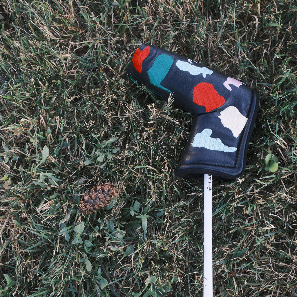 A golf club headcover with a colorful abstract pattern, fitted on a golf club shaft lying on a grassy ground next to a pinecone.