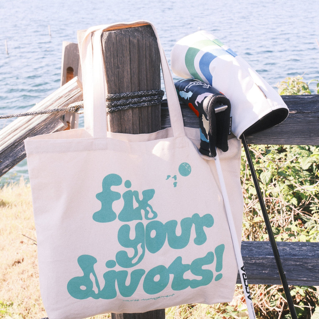 A cream canvas tote bag with teal text reading 'fix your divots!' hangs from a wooden post outdoors near water and foliage. Golf clubs also visible.