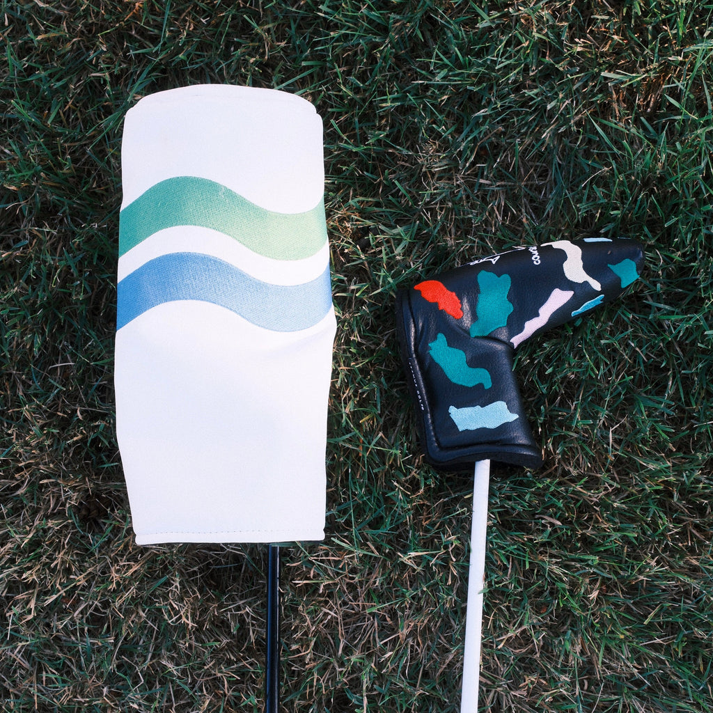 A golf club headcover set lying on grass, featuring a white headcover with blue and green wavy stripes and a black golf course-patterned putter headcover.