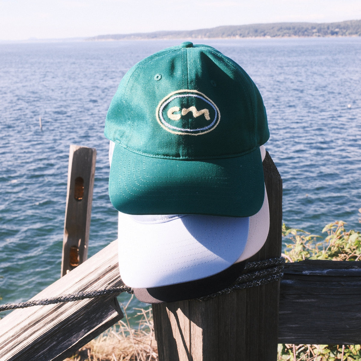 A green baseball cap with a white chain stitched circular logo sits on a dark wooden post outdoors, with water and shoreline visible in the background.