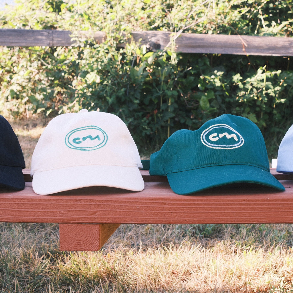 Four baseball caps in black, cream, teal, and light blue are lined up on a wooden bench outdoors with a rustic fence and greenery in the background. Each cap features a circular logo with the letters 'CM'.