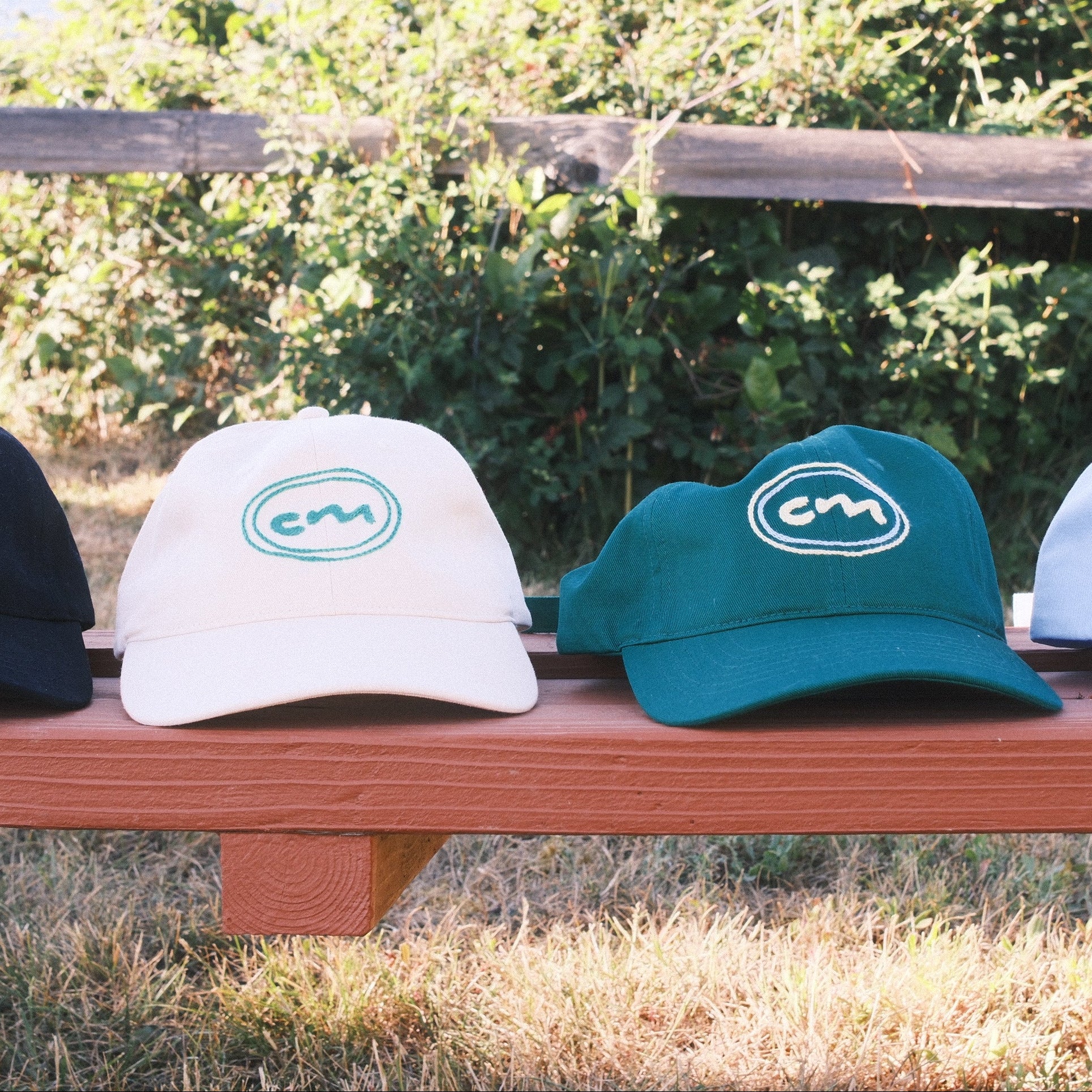 Four baseball caps in black, cream, teal, and light blue are lined up on a wooden bench outdoors with a rustic fence and greenery in the background. Each cap features a circular logo with the letters 'CM'.