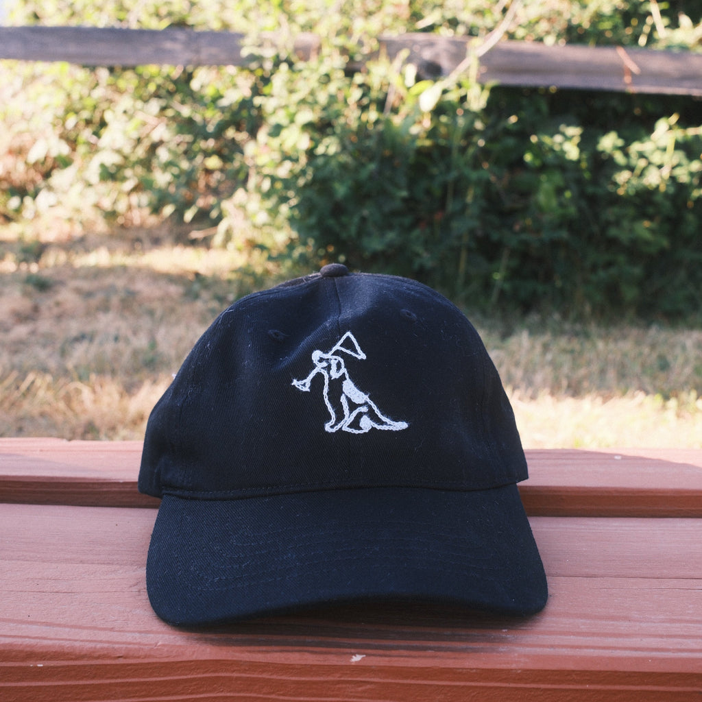 A black baseball cap with a white embroidered logo, resting on a reddish-brown wooden deck railing with a blurred outdoor background of trees and grass.