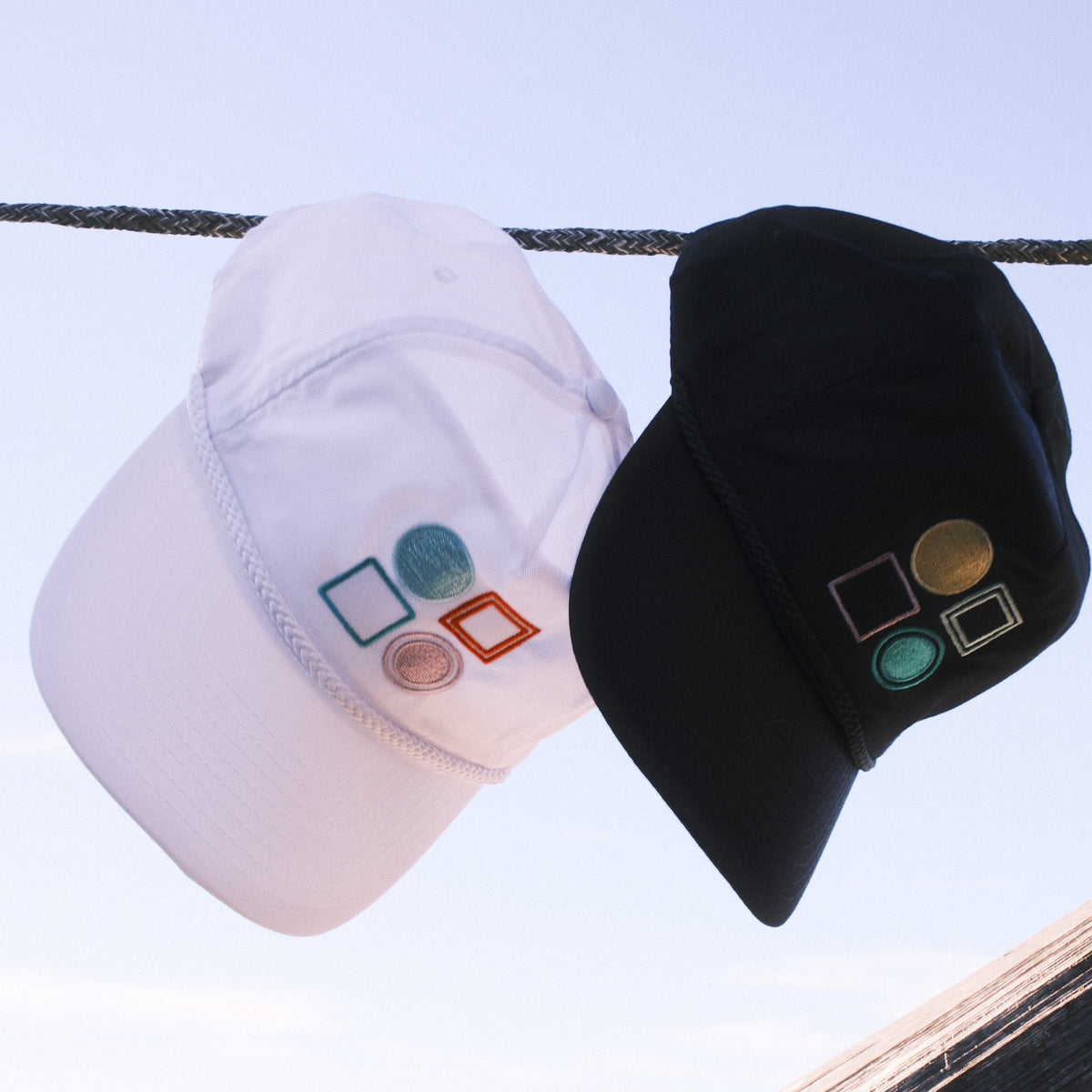Two baseball caps hanging outdoors: a black cap on top with embroidered geometric shapes, and a white cap below with matching embroidered shapes. The background shows a bright outdoor scene with a wooden structure and light sky.