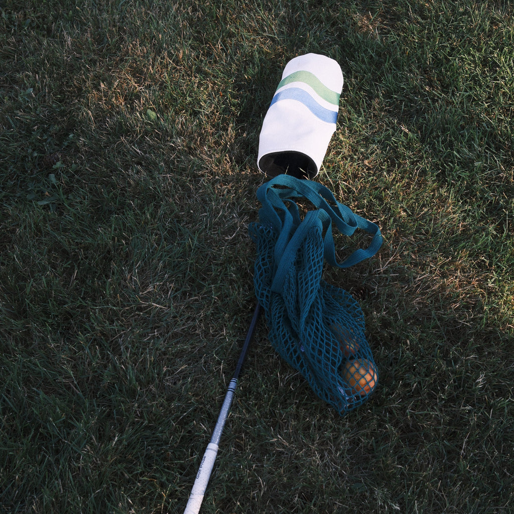 A white and blue striped golf club with head cover lies on its side on grass next to a teal mesh ball bag photographed outdoors on a grassy field.