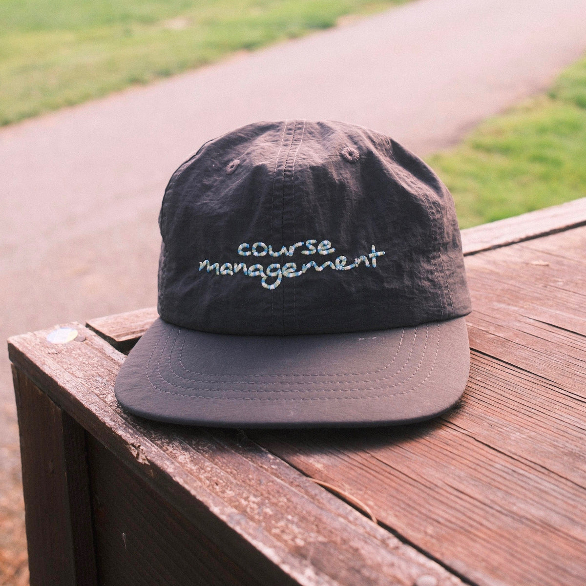 A dark gray flat-brim cap with 'course management' embroidered script on the front, resting on a wooden table outdoors with a blurred golf course background.