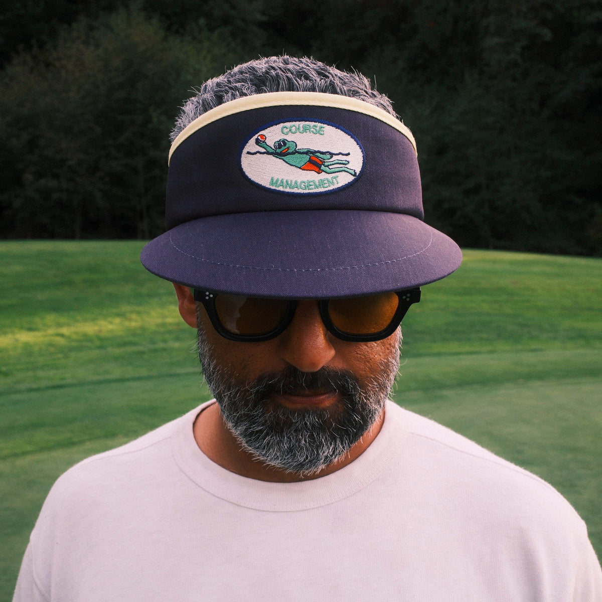 A man wearing a navy blue visor with a golf-themed patch and sunglasses, standing on a golf course green with a flag in the background.