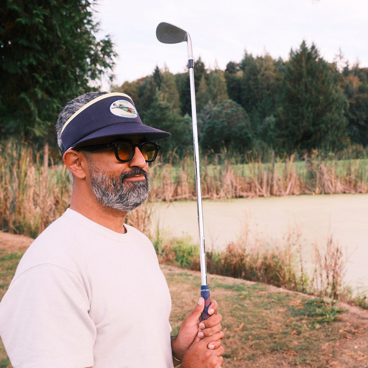 A man on a golf course holds a golf club, wearing a navy blue visor with a white logo, yellow-tinted sunglasses, a white t-shirt, and a gray beard. Trees and a pond are visible in the background.