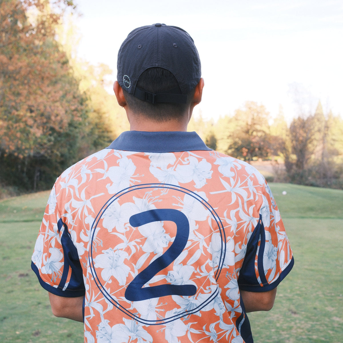 A man viewed from behind wearing a short-sleeve athletic jersey with a white, orange, and navy floral pattern, featuring the number 2 on the back. He is standing on a grassy field with trees in the background.
