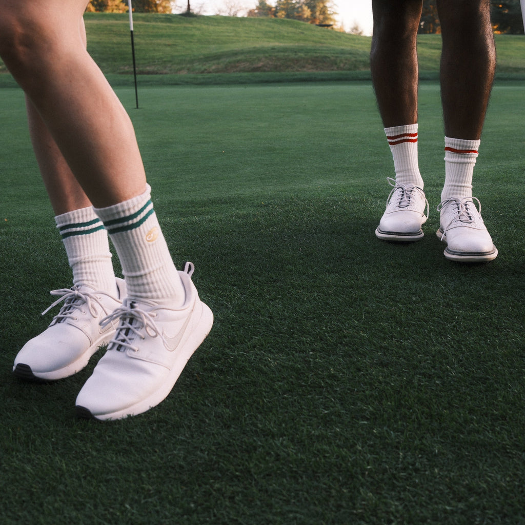 White athletic golf shoes worn by two golfers on a green golf course, with white socks and golf clubs visible.