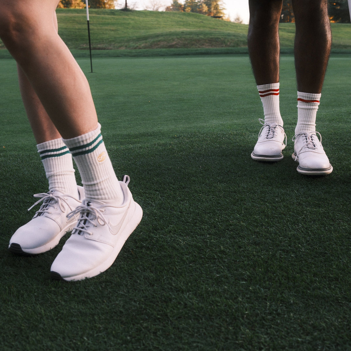 White athletic golf shoes worn by two golfers on a green golf course, with white socks and golf clubs visible.