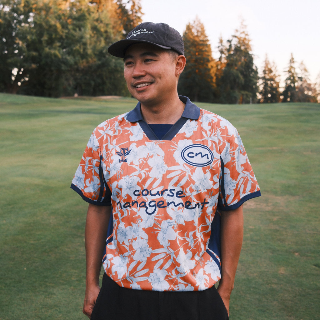 A man wearing a patterned orange and white golf course management jersey, black pants, and a black cap stands on a golf course lawn with trees in the background.