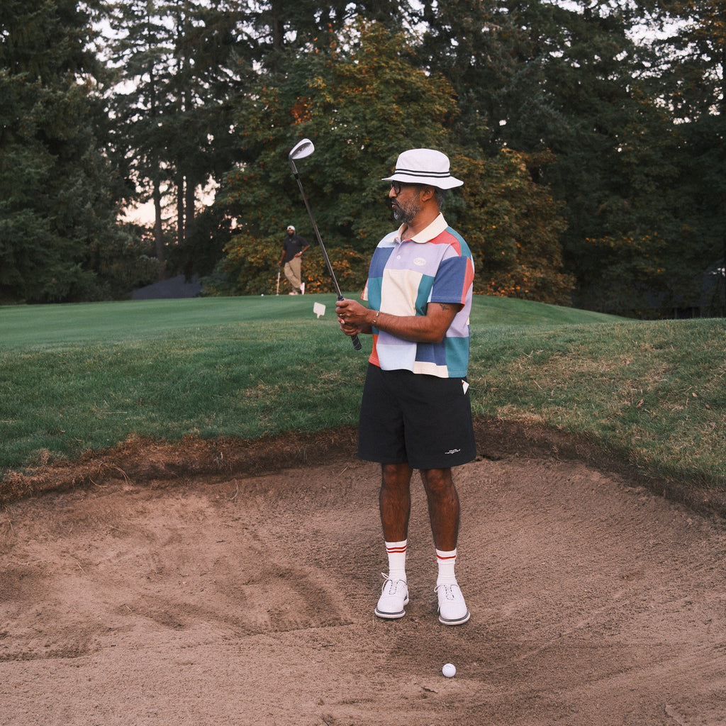 A man wearing a colorful geometric patterned polo shirt, black shorts, white socks, and a white hat stands in a sand bunker on a golf course, holding a golf club.