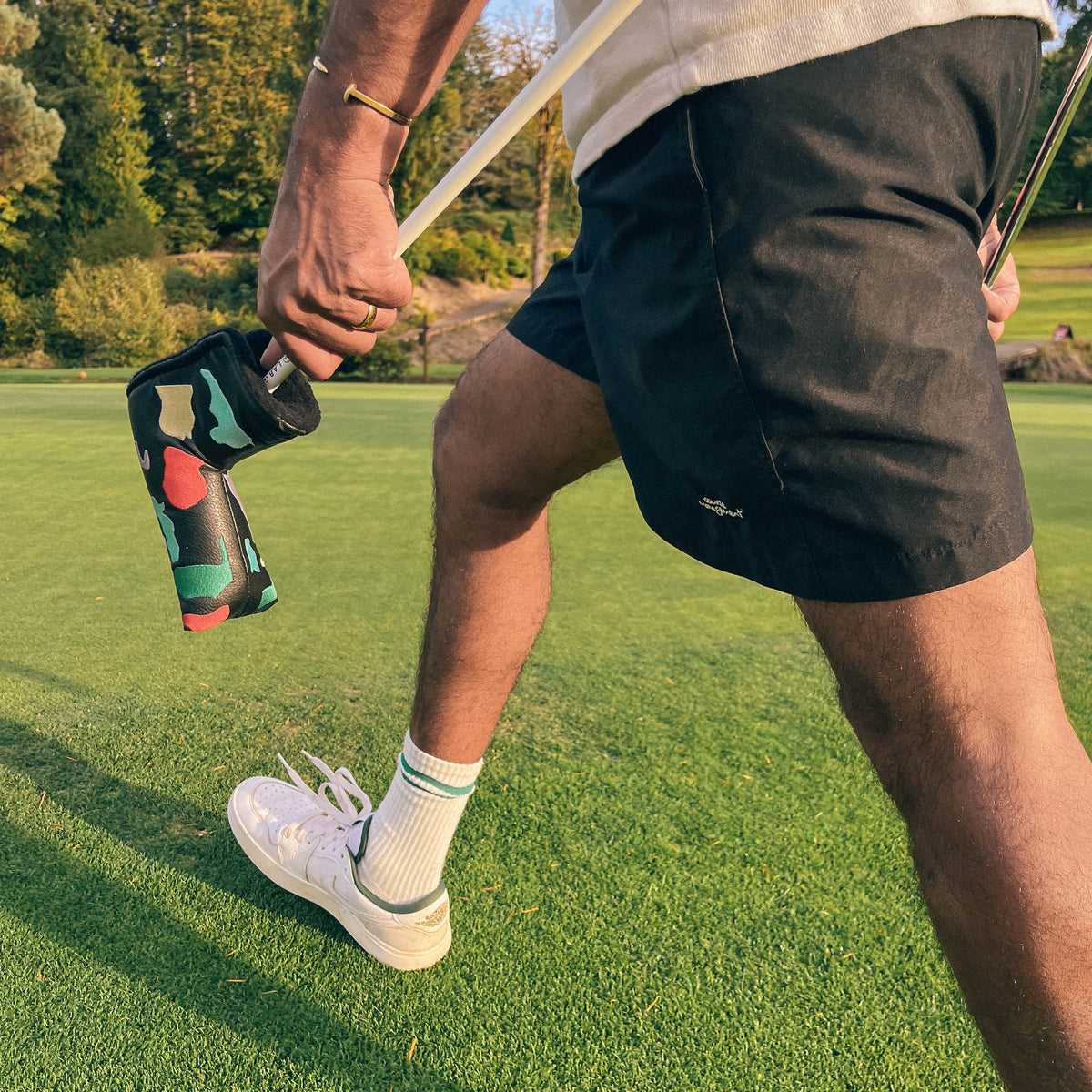 A close-up lifestyle shot of a person on a golf course wearing black shorts, white socks, and white golf shoes, holding a golf club with a colorful patterned headcover.