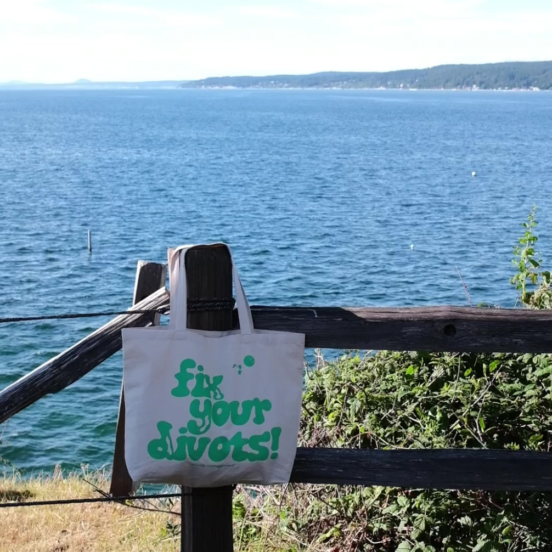 Tote bag displaying the text Fix Your Divots hangs on a fence post in front of the ocean.
